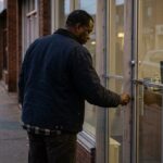 A small storefront office at dusk, closing time, one person locking the front door of a modest commercial unit on a quiet Canadian street, a stack of pa...